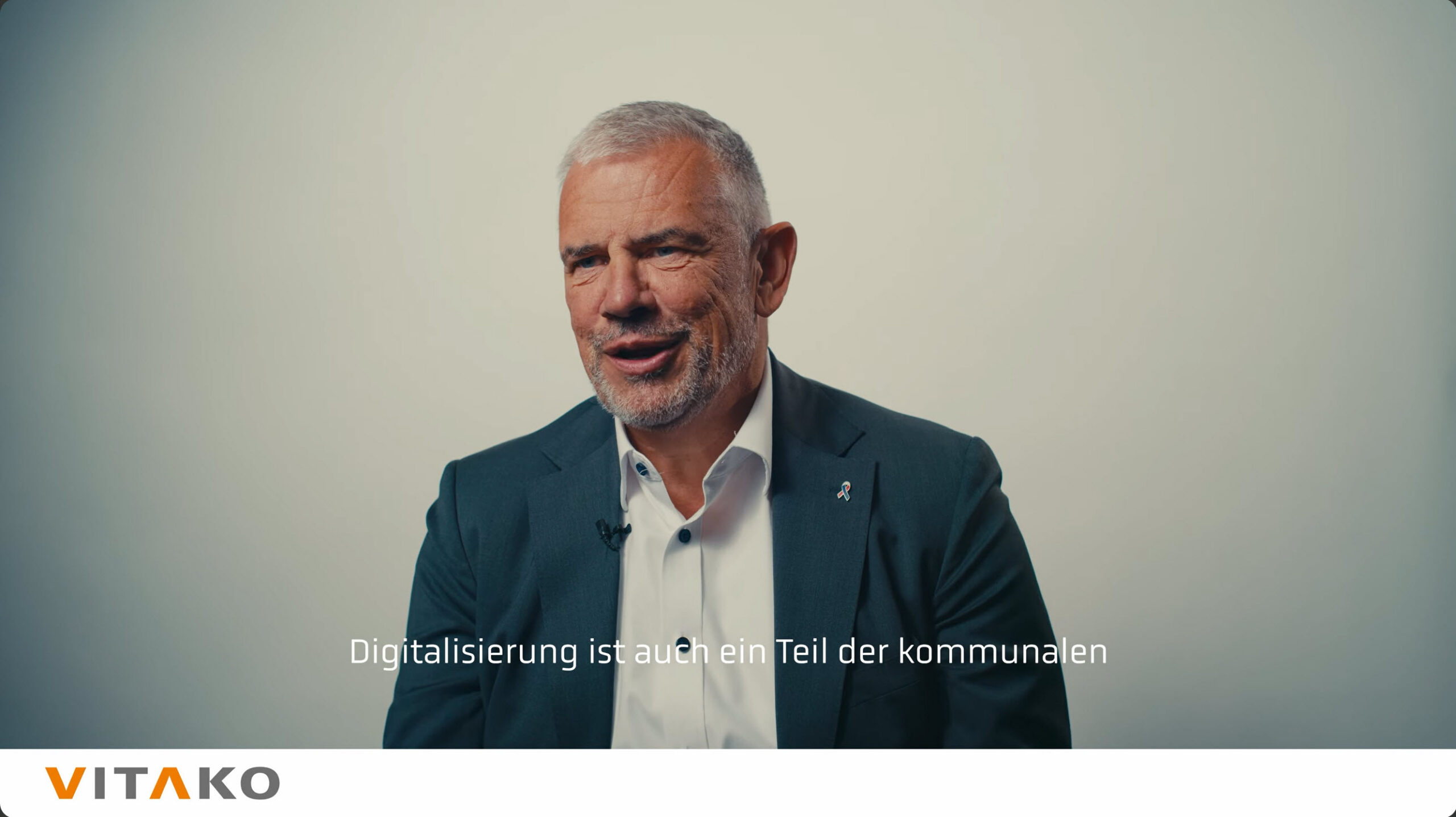 Screenshot: Mann im dunklen Anzug vor professionell ausgeleuchteter heller Wand