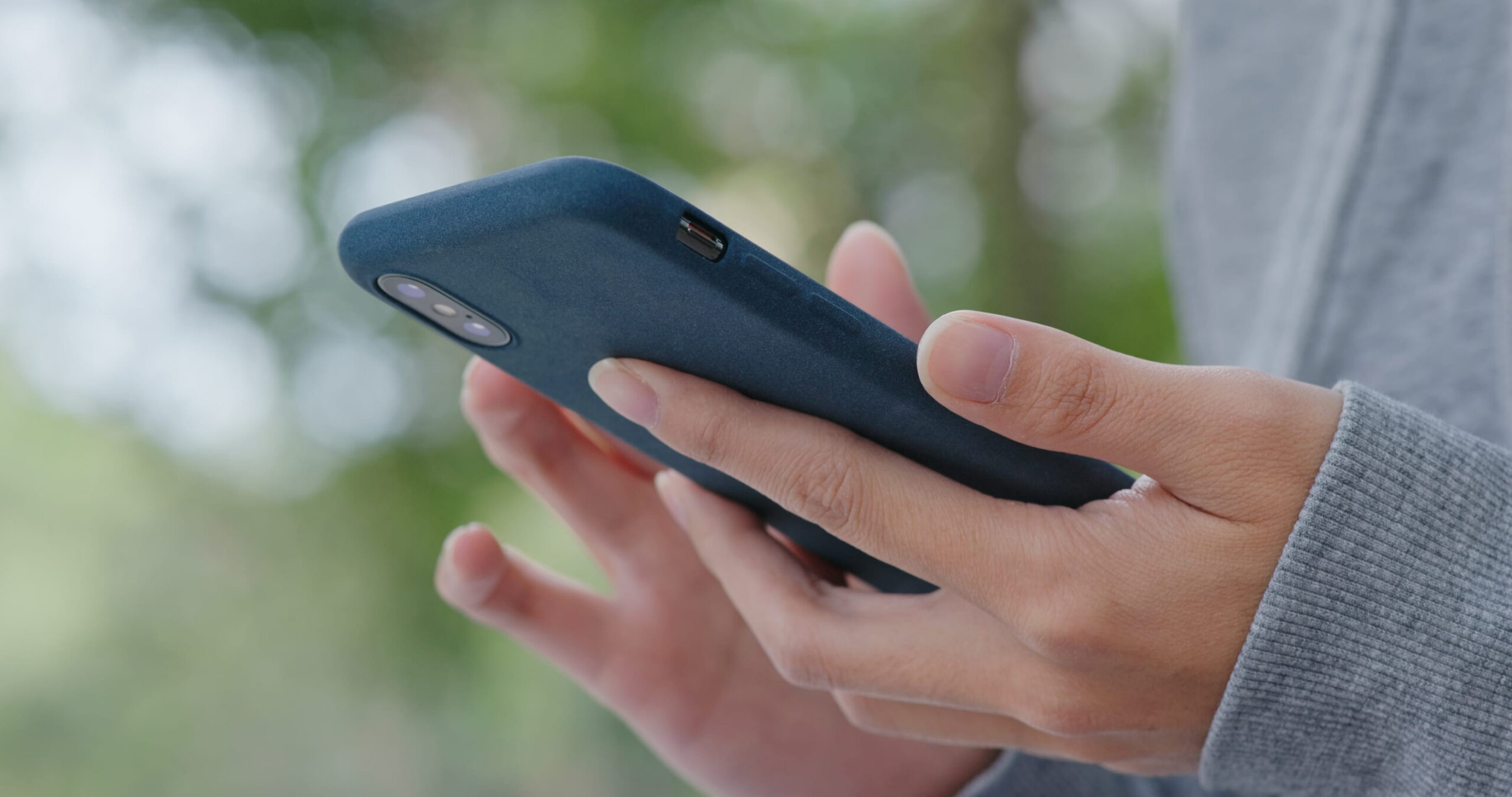 Nahaufnahme auf ein Paar Hände mit Smartphone, im Hintergrund unscharfe Vegetation