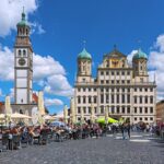 Blick über den belebten Augsburger Rathausplatz mit Außengastronomie auf das historische Rathaus und den Perlachturm vor blauem Himmel.