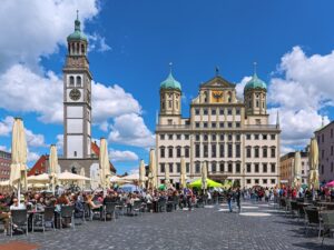 Blick über den belebten Augsburger Rathausplatz mit Außengastronomie auf das historische Rathaus und den Perlachturm vor blauem Himmel.