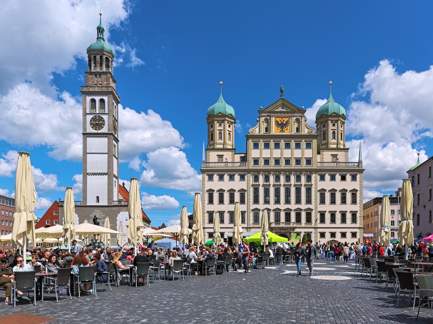 Blick über den belebten Augsburger Rathausplatz mit Außengastronomie auf das historische Rathaus und den Perlachturm vor blauem Himmel.