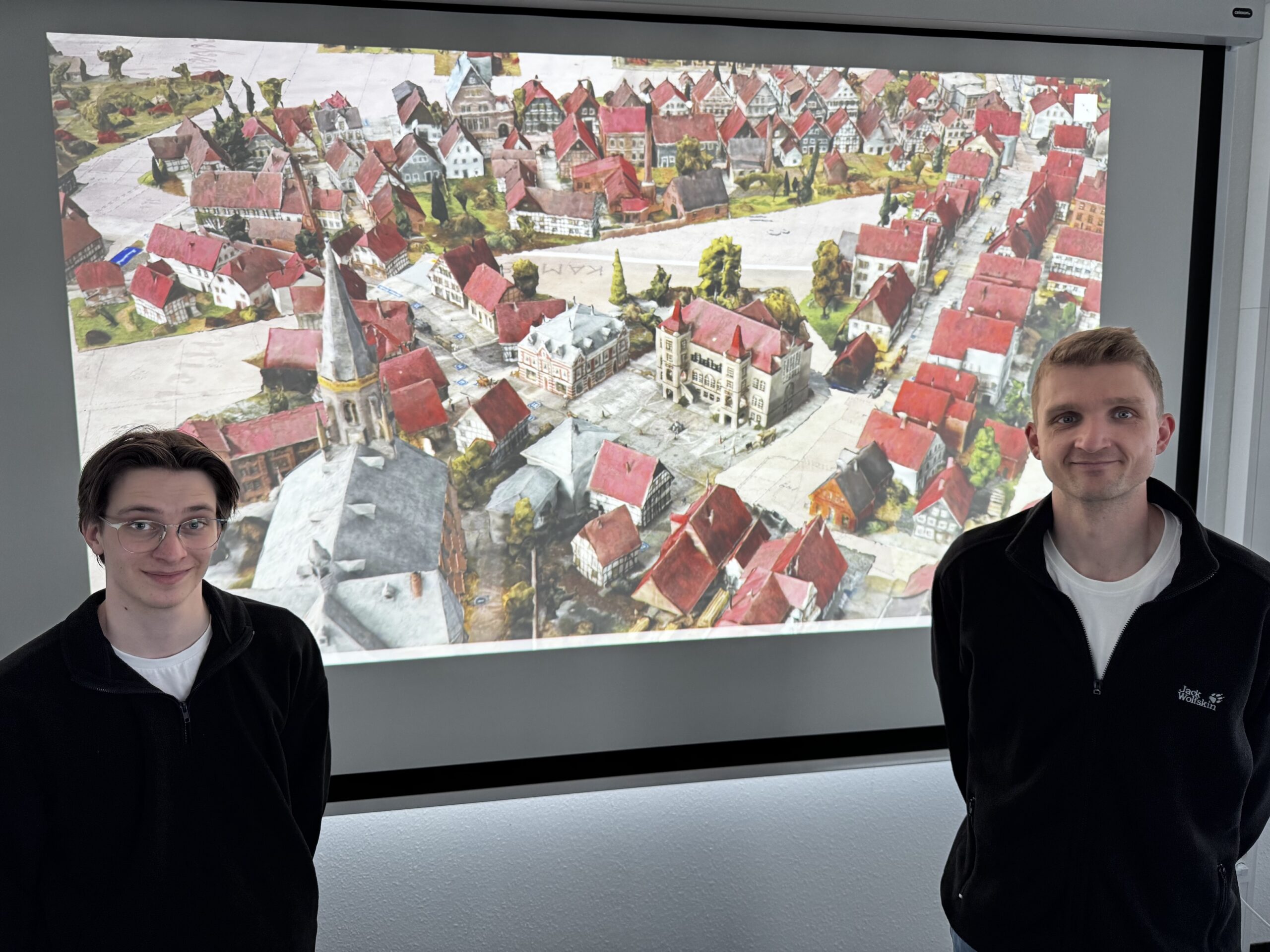 Zwei Personen vor einem Monitor auf dem historische 3D-Modell Gütersloh um 1900 zu sehen ist