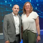 Ein Mann und eine Frau stehen für ein gemeinsames Foto vor einer blauen Wand und schauen in die Kamera.