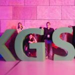 Große , dreidimensionale Buchstaben "KGST" vor einer rosa beleuchteten Wand, dahinter und dazwischen stehen Menschen