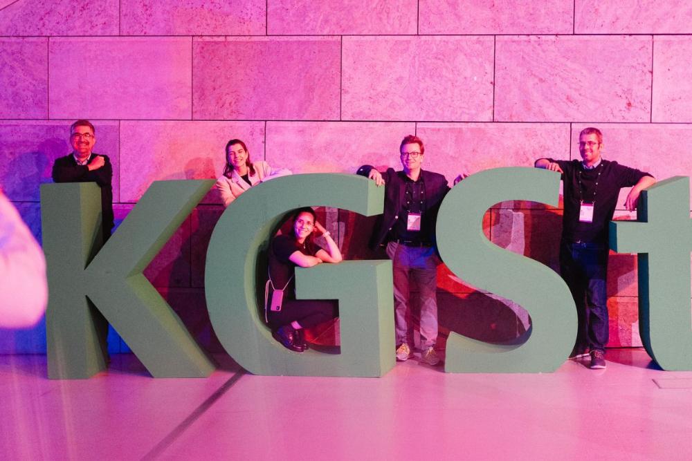 Große , dreidimensionale Buchstaben "KGST" vor einer rosa beleuchteten Wand, dahinter und dazwischen stehen Menschen