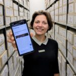 Junge Frau zwischen Archivregalen streckt ihr Handy Richtung Kamera
