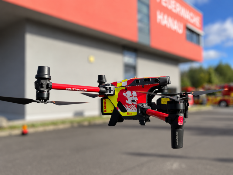Drohne in der Luft, im Hintergrund ist verschwommen ein Gebäude der Feuerwehr Hanau zu sehen
