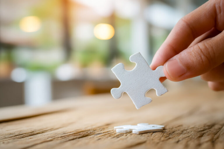 Eine Hand hält ein weißes Puzzlestück über einen Holztisch, auf dem ein weiteres Puzzleteil liegt